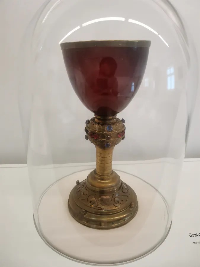 Ein kunstvoller Hoheitspokal aus Metall, umgeben von einer Glaskuppel, steht auf einer weißen Oberfläche. Der Pokal hat eine rot gefärbte Innenfläche und ist auf einem verzierten Sockel mit bunten Steinen montiert.