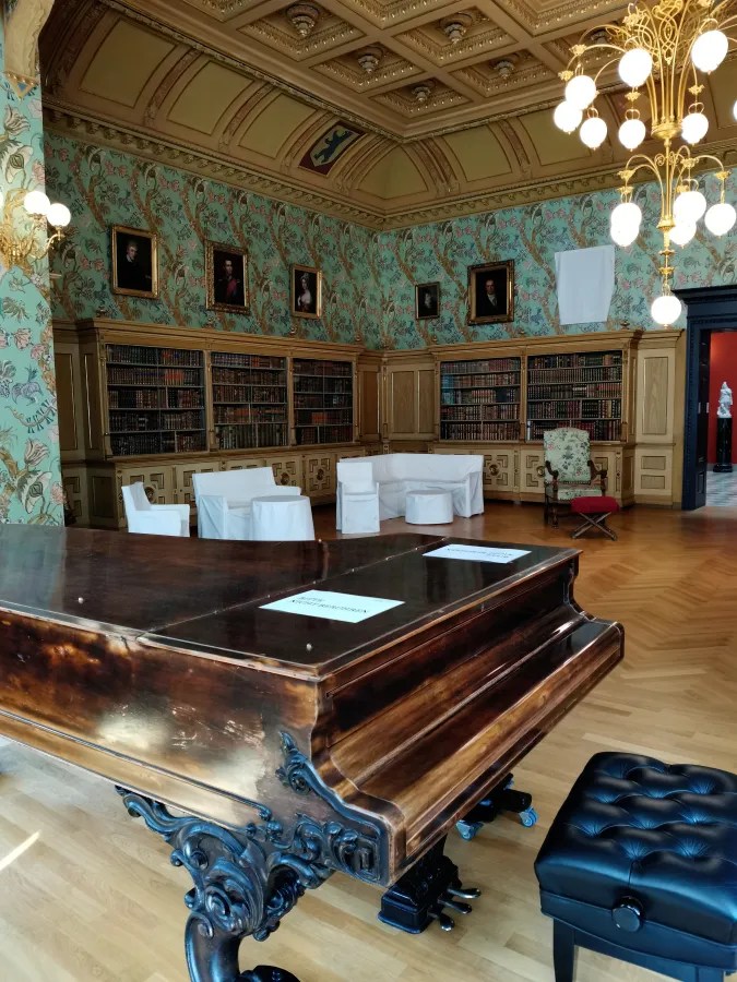 Ein elegantes Musikzimmer mit einem historischen Klavier, umgeben von Bücherregalen und kunstvoll gestalteten Wänden.