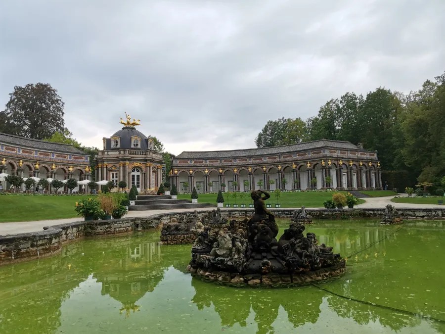Ein barockes Gebäude mit goldenen Akzenten umgeben von einem grünen Garten und einem Teich mit einer Statue in der Mitte.