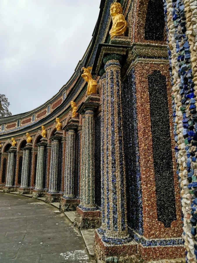 Detailaufnahme der kunstvoll dekorierten Fassade mit bunten Steinen und goldenen Skulpturen in einem Garten.