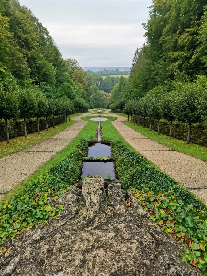 Symmetrischer Garten mit grünen Alleen, einem Wasserlauf und Bäumen, umgeben von Wald, an einem bewölkten Tag.