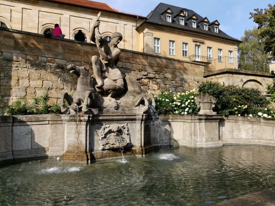 Bronze-Wasserspiel mit Figuren und Tieren in einem Garten, umgeben von gepflanzten Blumen und einem historischen Gebäude im Hintergrund.