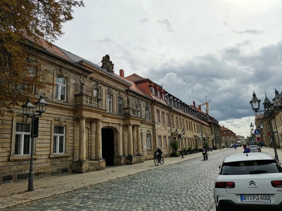 Blick auf eine historische Straße mit Statuen an den Gebäuden und einem grauen Himmel im Hintergrund.