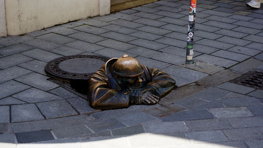 Bronzestatue eines Kanalarbeiters, der aus einem Schacht herausblickt, umgeben von gepflastertem Boden und einem Straßenpoller.