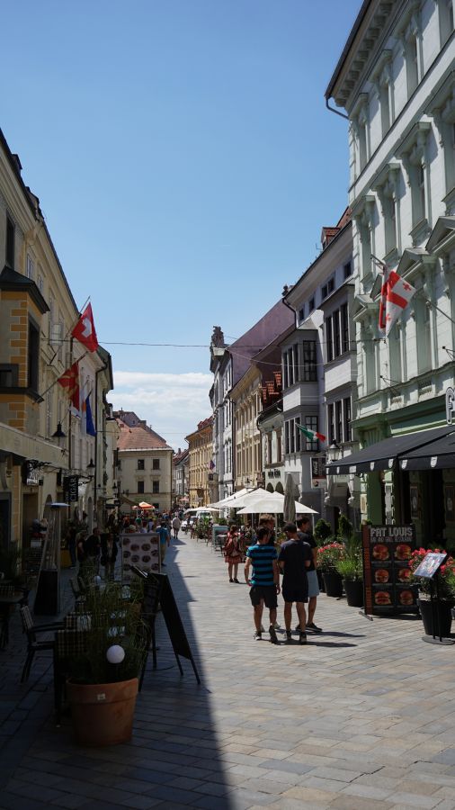 Blick auf eine belebte Straße in Bratislava mit Cafés, Geschäften und Menschen, umgeben von historischen Gebäuden und blauem Himmel.