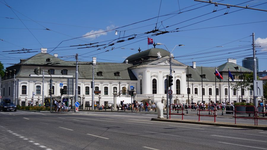 Historisches Gebäude in Bratislava mit Menschenschlange vor dem Eingang und Oberleitungen im Vordergrund.