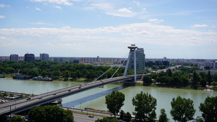 Panoramablick auf die Donau mit einer modernen Brücke, umgeben von grünen Bäumen und städtischer Architektur in Bratislava unter blauem Himmel.