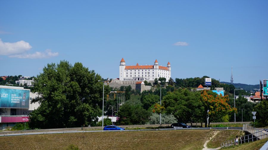 Blick auf die Burg Bratislava, umgeben von grünen Bäumen und einem blauen Himmel, mit modernen Gebäuden im Vordergrund.