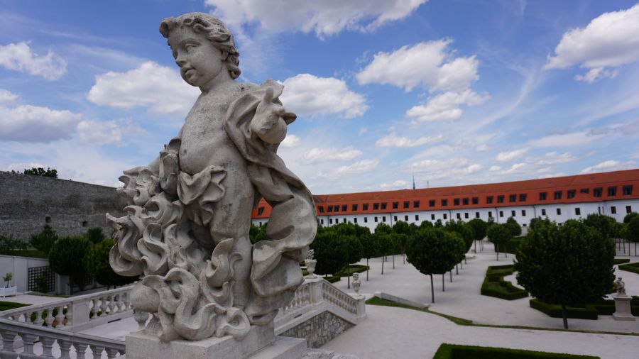 Eine Statue eines Engels in einem Garten mit symmetrischen Hecken und Wolken am blauen Himmel im Hintergrund.