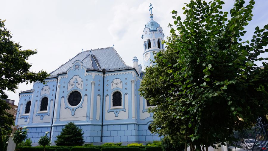 Himmelblaue Jugendstilkirche in Bratislava, umgeben von Bäumen und einem grünen Garten.