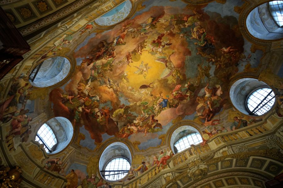 Decke mit farbenfrohem Fresko in der Hofburg, zeigt Szenen von Engeln und mythologischen Figuren unter einem hellen Himmel.
