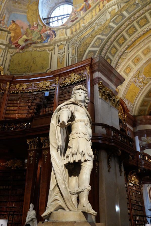 Eine Statue eines Kriegers in einer prächtigen Bibliothek mit goldenen Verzierungen und hohen Bücherregalen im Hintergrund.
