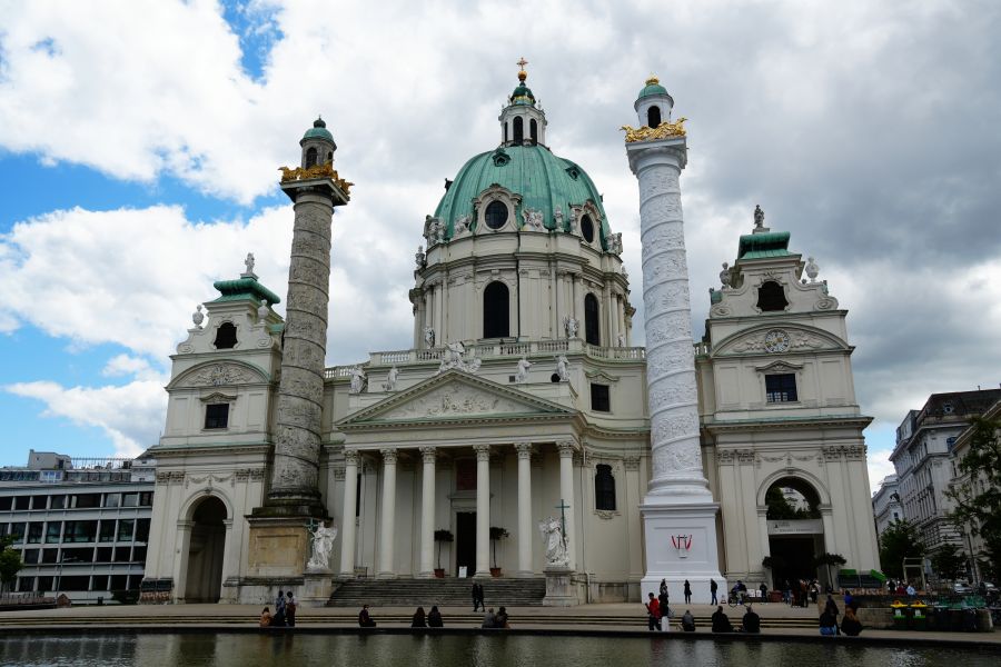 Blick auf die Karlskirche in Wien mit ihrer markanten grünen Kuppel, flankiert von zwei Säulen und einem Wasserbecken im Vordergrund, unter einem bewölkten Himmel.