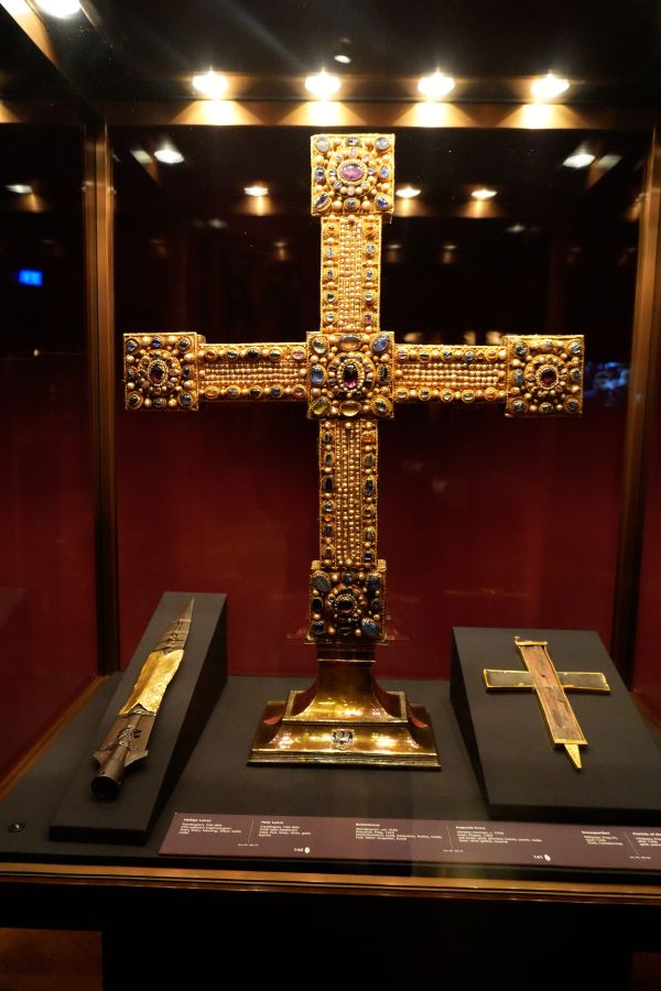 Goldener Kreuz mit Edelsteinen, ausgestellt in einer Vitrine, umgeben von weiteren kunstvoll gestalteten Objekten.
