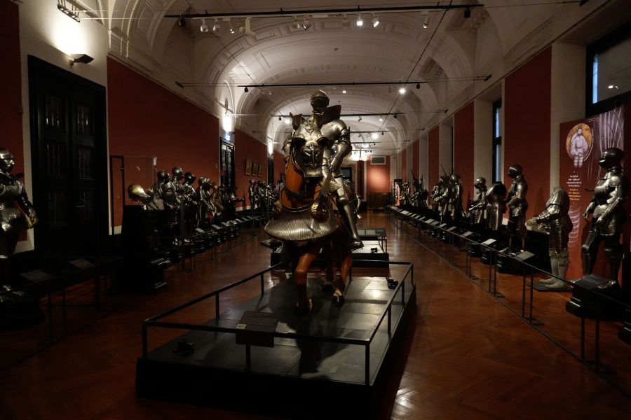 Eine Ausstellung in der Hofburg zeigt ritterliche Rüstungsteile und Figuren, die auf einem Pferd sitzen, umgeben von weiteren Rüstungen in einer eleganten Halle.
