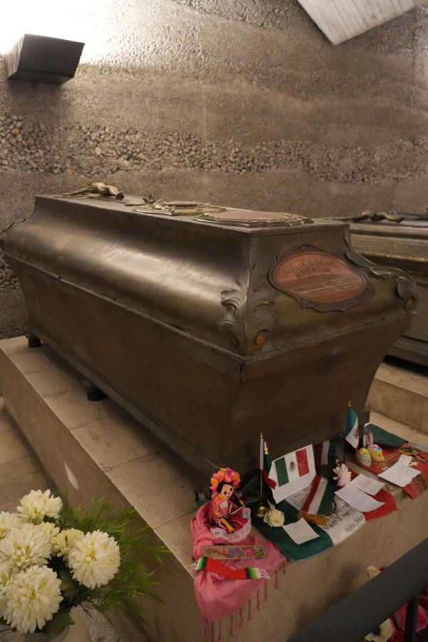 Ein bronzener Sarg mit Gedenkgegenständen und Blumen in einer Gruft, umgeben von Steinen.