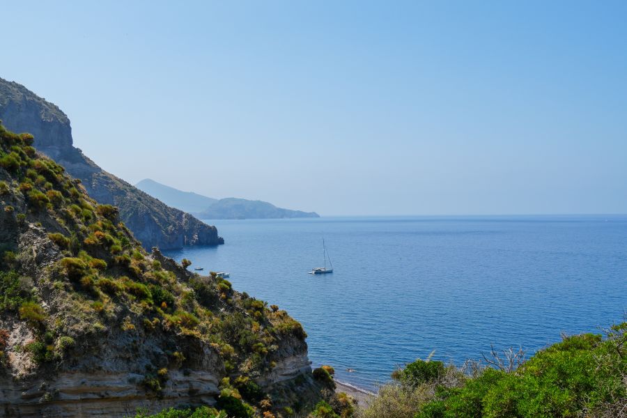 Blick auf das glasklare Wasser vor der Küste, umgeben von Hügeln und Bergen. Ein Segelboot schwimmt ruhig auf dem Meer, während die klare Himmel blaut.