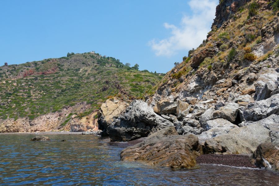Blick auf eine Küstenlandschaft mit steilen Klippen, umgeben von klarem Wasser und grün bewachsenen Hügeln unter einem blauen Himmel.