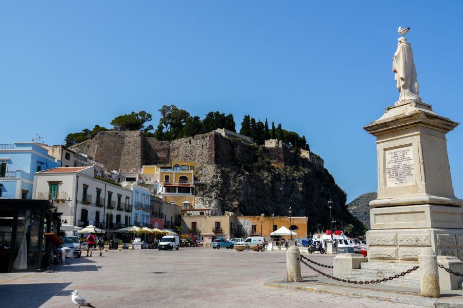 Ein Blick auf den Hafen von Lipari mit bunten Gebäuden und einer Statue im Vordergrund, während im Hintergrund eine Festungsmauer sichtbar ist.