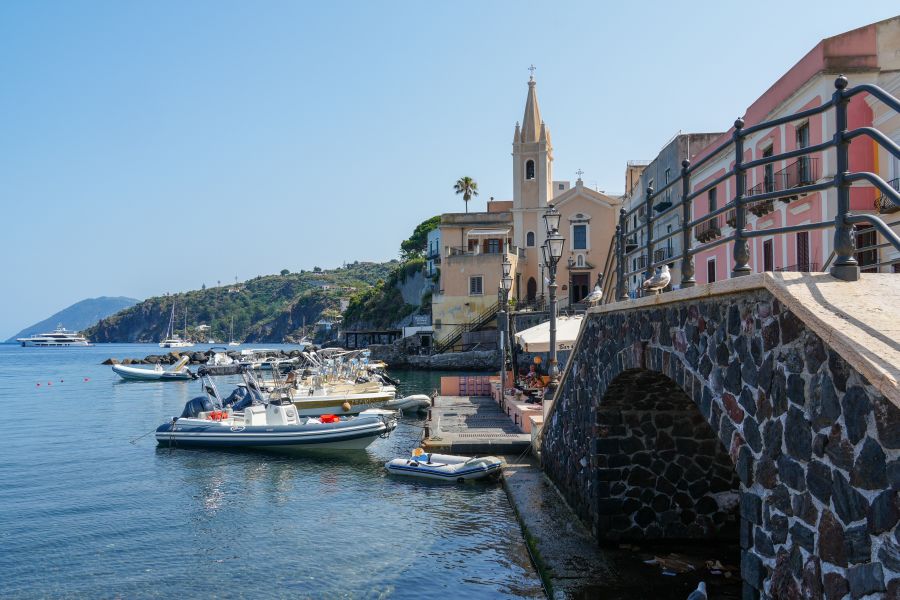 Blick auf einen malerischen Hafen mit Booten und einer Kirche im Hintergrund, umgeben von felsiger Küste und mediterranen Häusern.