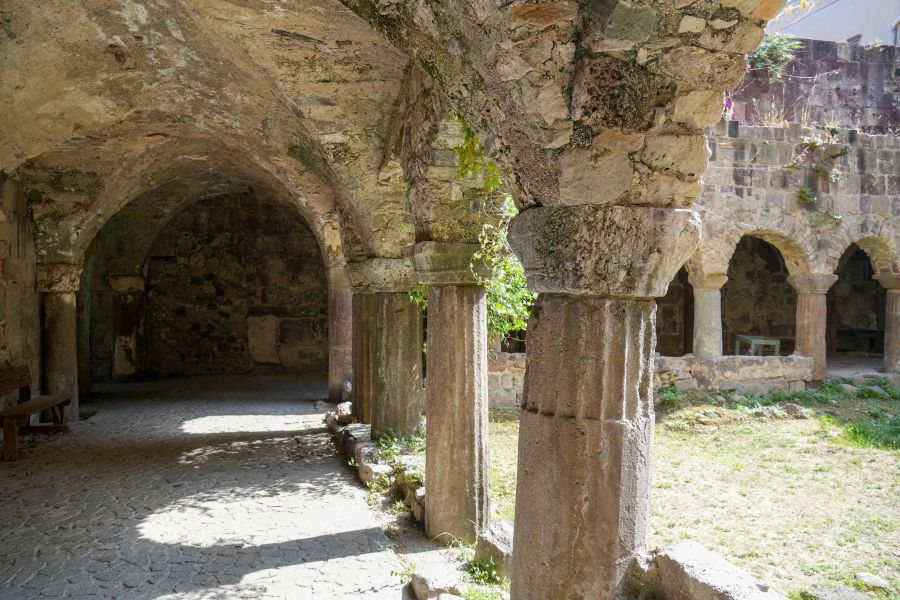 Blick auf einen alten, steinernen Innenhof mit Bögen und Säulen, umgeben von einer kleinen, grünen Wiese.