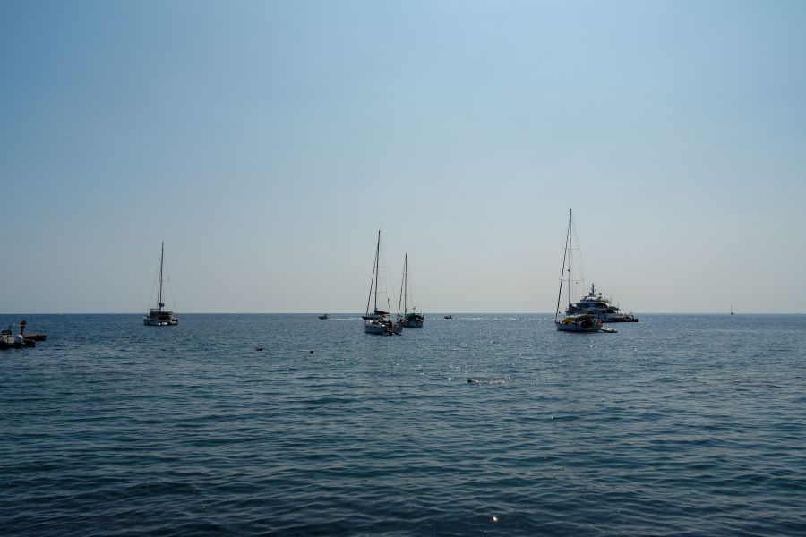 Blick auf mehrere Segelboote, die ruhig auf dem glitzernden Wasser des Mittelmeers vor einem klaren, blauen Himmel liegen.