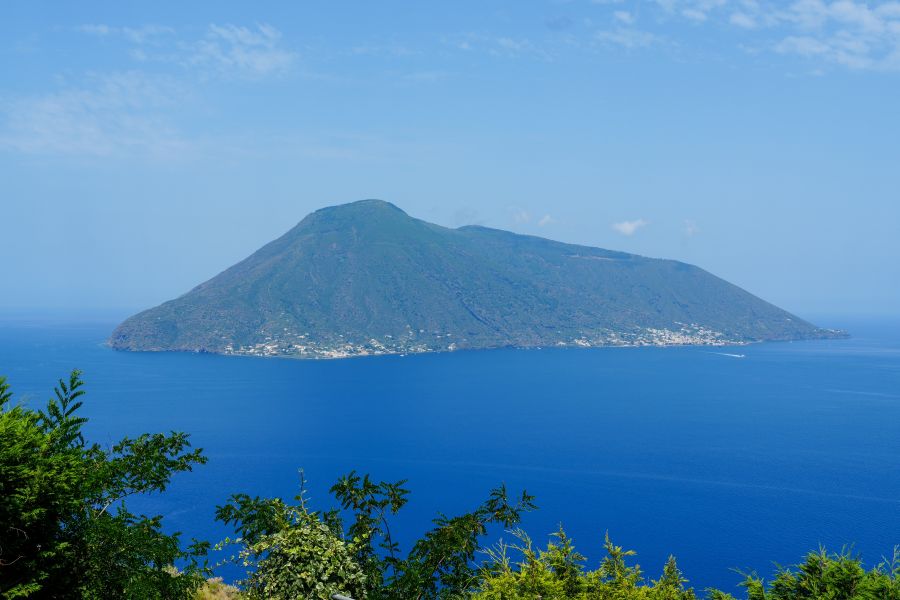 Blick auf die vulkanische Insel Salina im strahlend blauen Meer, umgeben von grünem Bewuchs im Vordergrund.