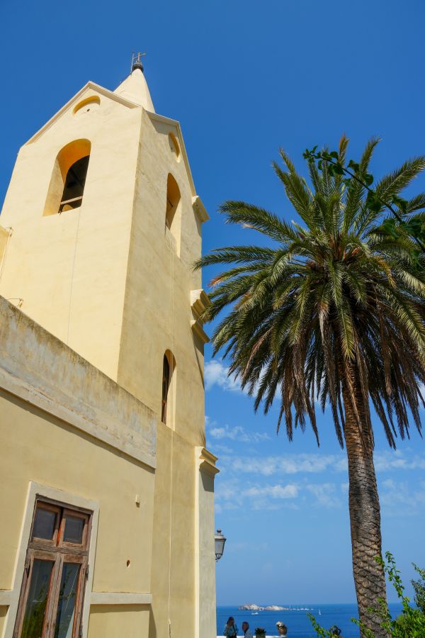Ein gelbes Gebäude mit Kirchturm und einer Palme im Vordergrund, vor einem strahlend blauen Himmel und einem Blick auf das Meer.