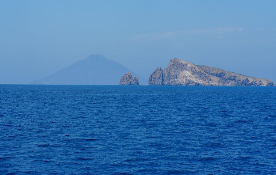 Blick auf die Liparischen Inseln mit dem Stromboli im Hintergrund und den felsigen Küsten im Vordergrund, umgeben von klarem blauem Wasser.