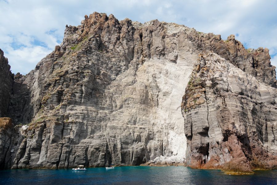 Aussicht auf steile, mächtige Klippen, die angrenzend an das glitzernde blaue Wasser der Liparischen Inseln zu sehen sind. Im Vordergrund sind zwei kleine Boote zu erkennen.