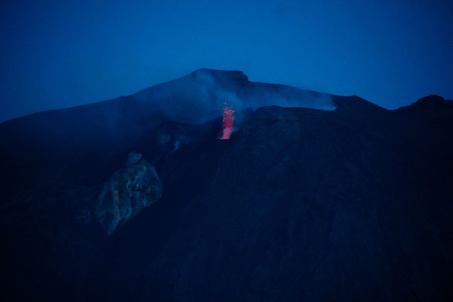Ein aktiver Vulkan (Stromboli) mit Lavaausbruch in der Dämmerung, umgeben von Rauch und einer dunklen Berglandschaft.