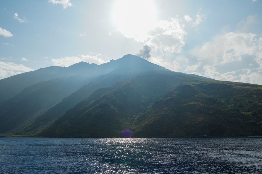 Der aktive Vulkan Stromboli mit einer Rauchsäule, die aus seiner Spitze emporsteigt, bei sonnigem Wetter und klarem Himmel, im Vordergrund das glitzernde Wasser des Mittelmeers.