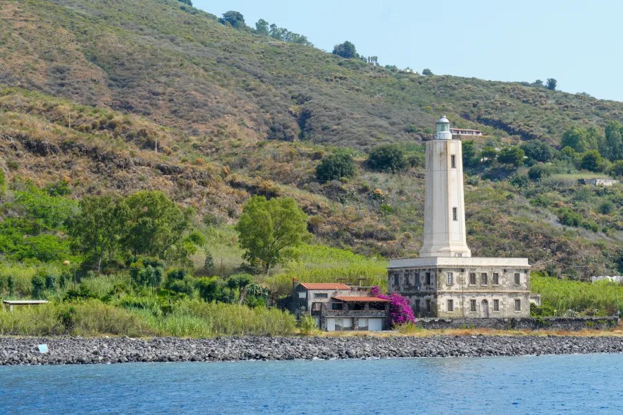 Ein Leuchtturm auf einer grünen Küstenlandschaft mit Hügeln im Hintergrund und blauem Wasser im Vordergrund.