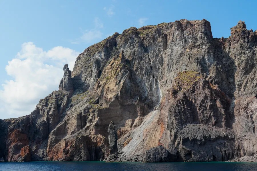 Blick auf eine steile, felsige Küste mit verschiedenen Farbtönen, darunter Braun, Grau und etwas Grün, unter strahlend blauem Himmel.