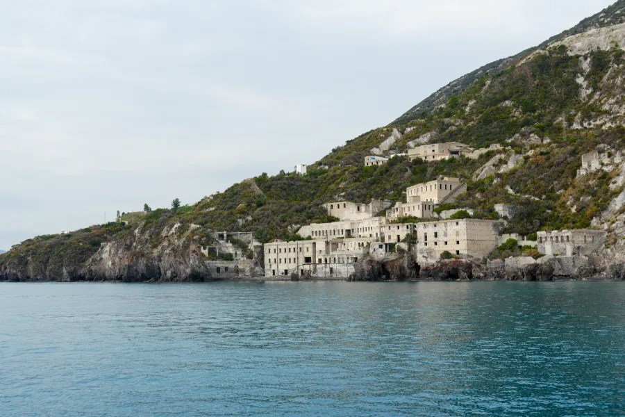 Panoramablick auf eine verlassene Gebäudegruppe an der Küste von Lipari, umgeben von hügeliger Landschaft und Wasser.