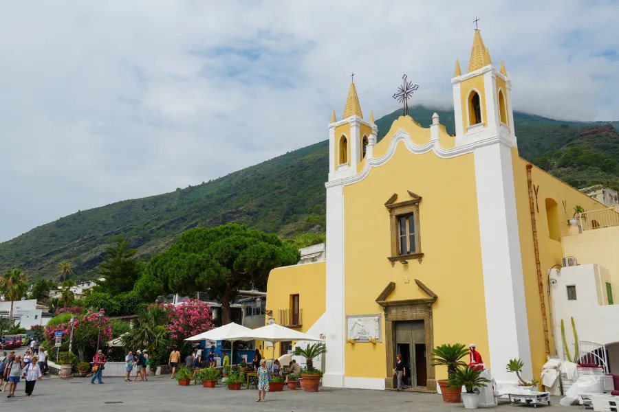 Blick auf eine gelbe Kirche mit zwei Türmen vor einer grünen Berglandschaft in Salina. Menschen gehen vor der Kirche entlang, umgeben von Pflanzen und Bäumen.