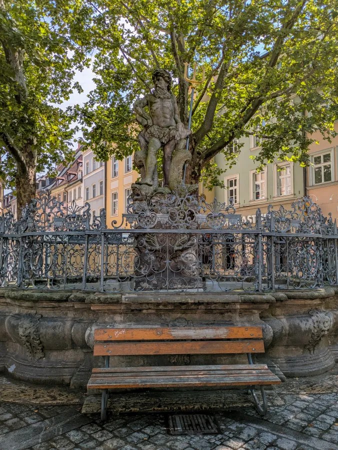 Neptun-Brunnen in Bamberg, Deutschland, mit einer Statue von Neptun und einer Holzbank im Vordergrund.