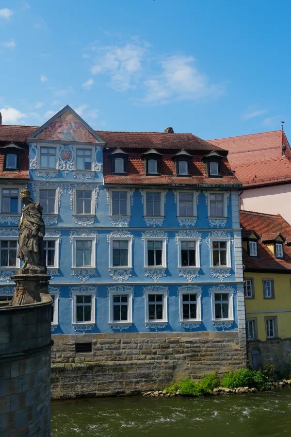 Blick auf ein historisches, blaues Gebäude mit bunten Wandmalereien direkt am Ufer der Regnitz in Bamberg, Deutschland. Eine Statue befindet sich im Vordergrund.