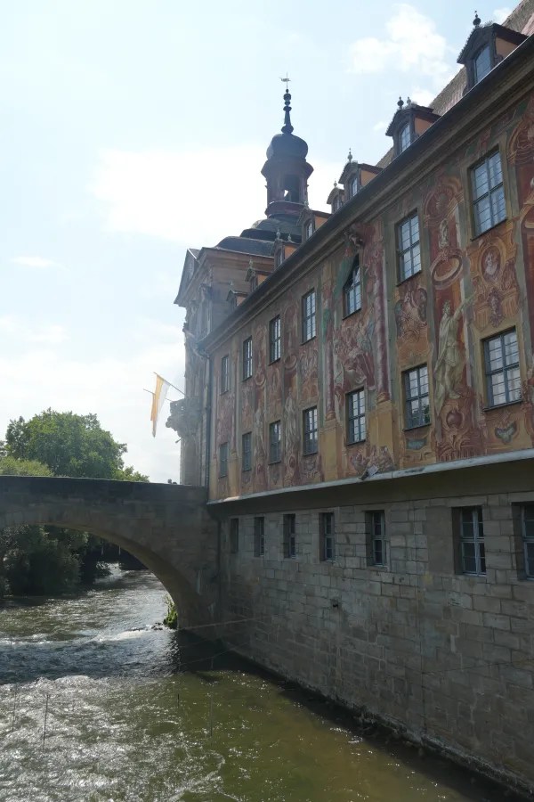 Das alte Rathaus in Bamberg, Deutschland, direkt am Wasser der Regnitz, mit bunten Wandmalereien und einer Brücke im Vordergrund.
