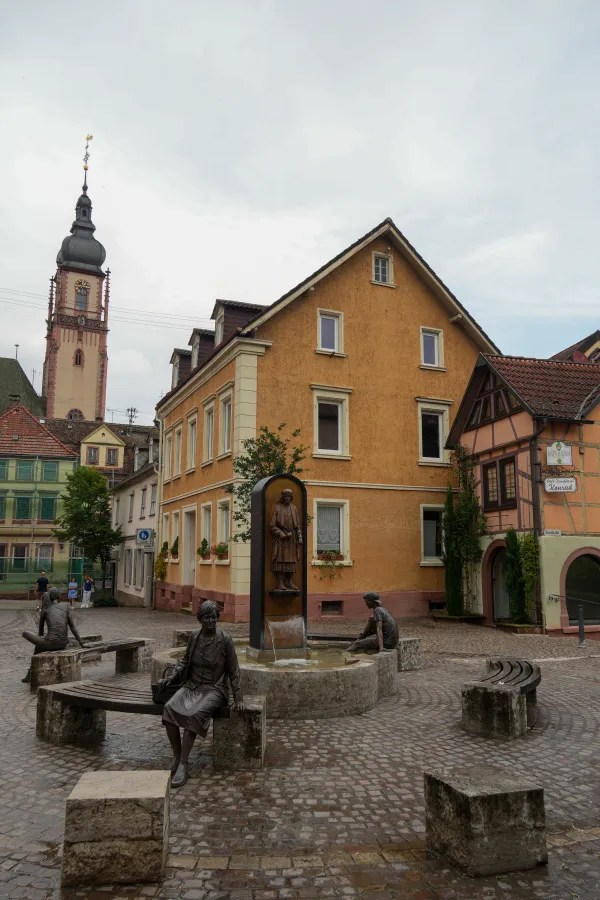 A town square featuring a fountain surrounded by benches and three bronze statues depicting people, with historic buildings and a church tower in the background.