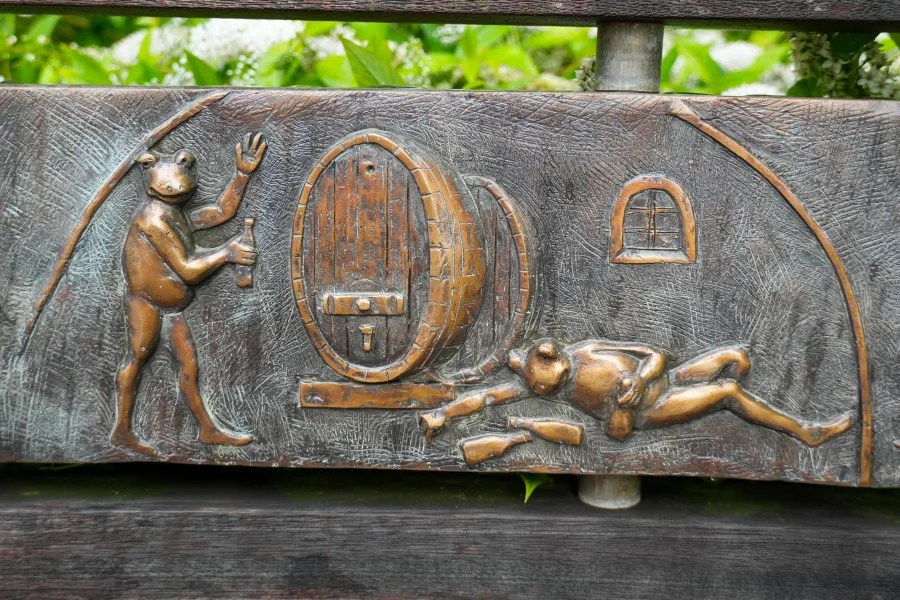 Bronze relief depicting a frog holding a bottle and a barrel, with a figure lying on the ground.