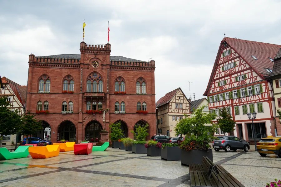 A colorful public square featuring the historic town hall with decorative windows and flags, surrounded by traditional half-timbered buildings and modern seating in various colors.