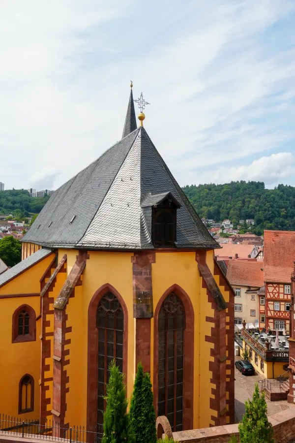 A close-up view of a church building featuring a pointed roof and large stained glass windows, with a green hillside in the background.