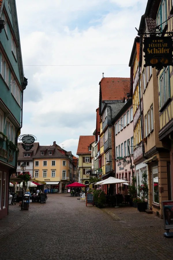 A picturesque street scene featuring colorful half-timbered houses, cafes with umbrellas, and a cloudy sky.
