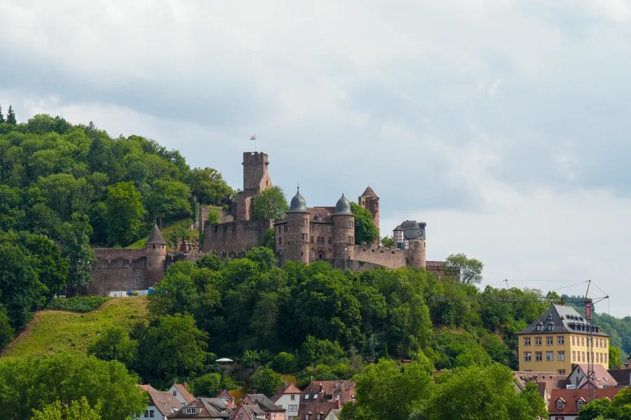 A view of a historic castle perched on a hill, surrounded by lush green trees and a small village at its base, under a cloudy sky.