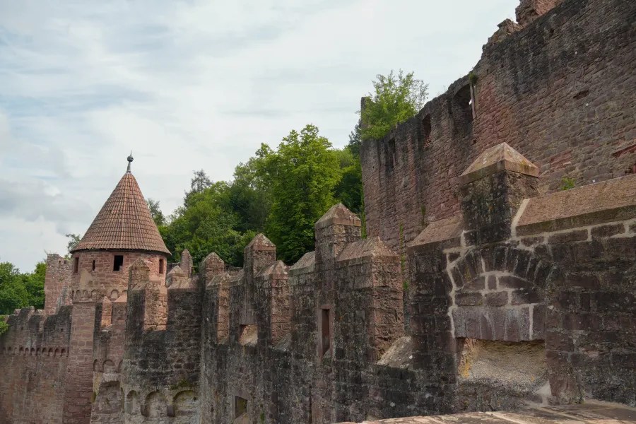 A view of the stone wall and tower of an old castle, partially surrounded by trees, with a cloudy sky in the background.