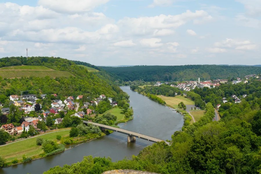 Panoramic view of a river winding through lush green hills and a small town, with a bridge crossing the water under a partly cloudy sky.