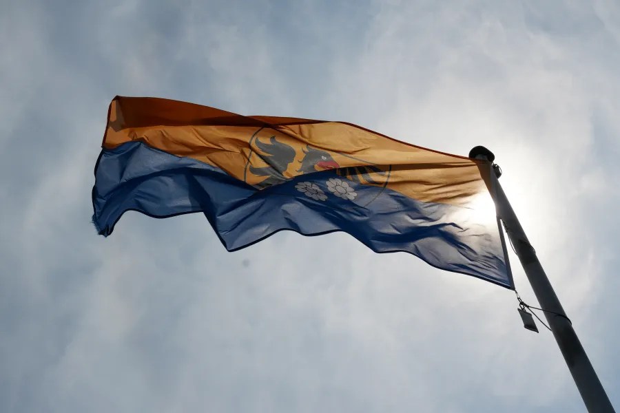 A flag featuring blue and orange colors, with a coat of arms, waving against a cloudy sky.