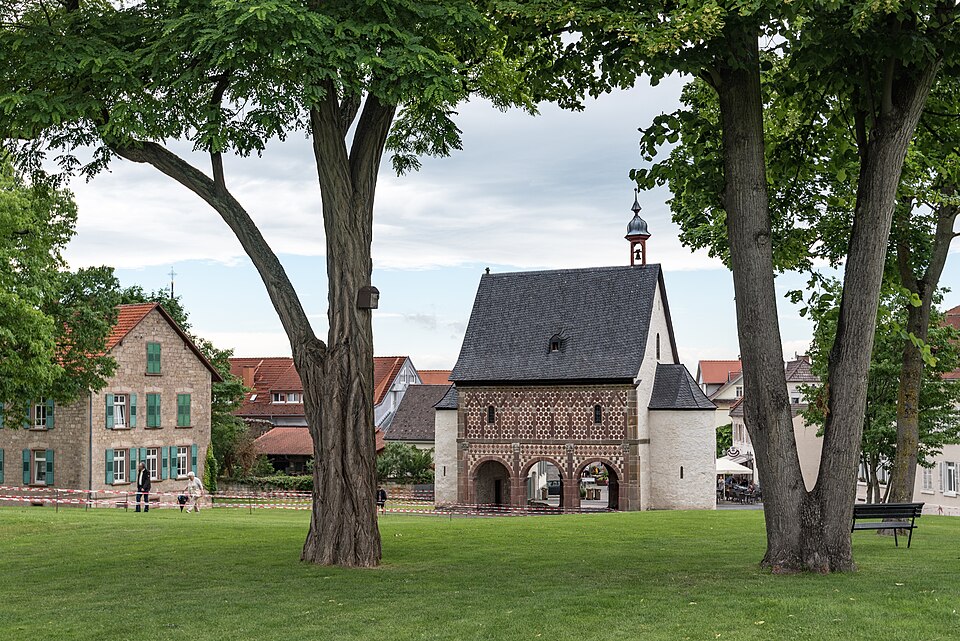 Blick auf ein historisches Gebäude mit Fensterläden in einem Park, umgeben von Bäumen und einer Wiese.
