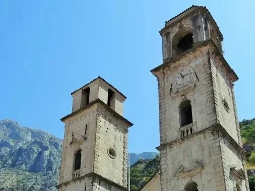 Zwei historische Kirchtürme mit detaillierter Architektur vor einem klaren blauen Himmel und Berglandschaft im Hintergrund.
